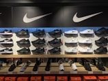 A display of various Nike shoes arranged on shelves, featuring both black and white sneakers. The shelves are backlit, highlighting the shoes, with the large Nike logo prominently displayed above. Price tags are visible on some shoes, and orange Nike shoe boxes are stacked below the display.