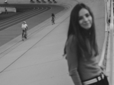 A black and white image featuring a blurred young woman in the foreground, standing near a cycling track. In the background, two cyclists can be seen riding on the track. The setting appears to be an outdoor sports venue.
