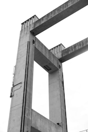 A concrete architectural structure with two large vertical towers connected by horizontal beams, likely part of a bridge or industrial installation. The material is weathered, showcasing a textured surface, and there are elements such as cables or pipes running along the sides.