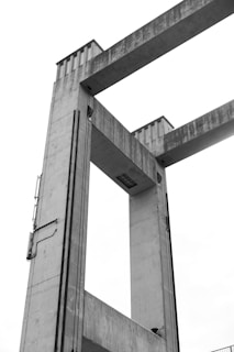 A concrete architectural structure with two large vertical towers connected by horizontal beams, likely part of a bridge or industrial installation. The material is weathered, showcasing a textured surface, and there are elements such as cables or pipes running along the sides.