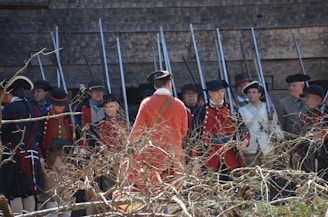 A group of people dressed in historical military uniforms, mostly red and blue, are standing in front of a wooden structure, holding rifles with bayonets. The scene includes leafless branches in the foreground.