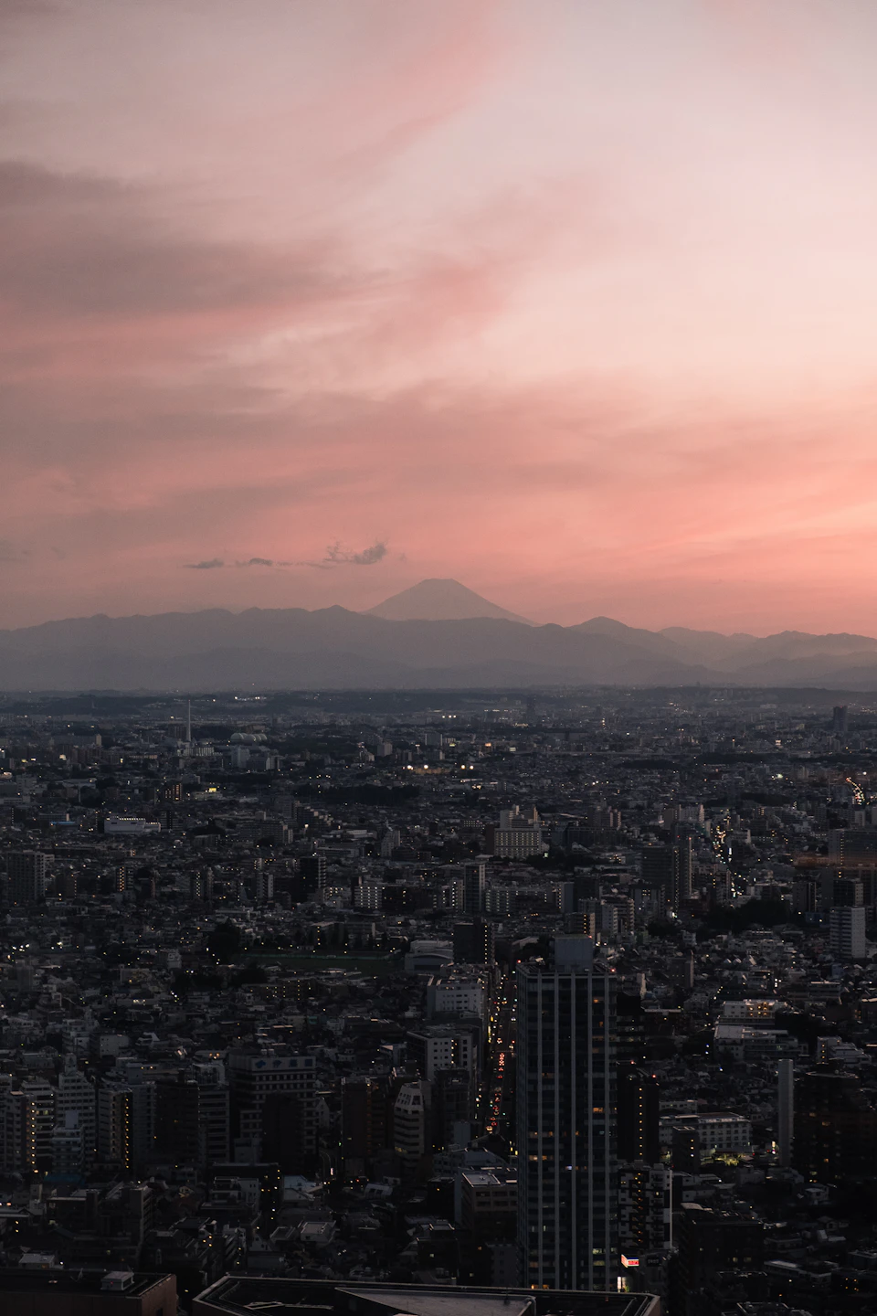 Cityscape at sunset with pink and orange hues, sun setting behind distant mountains
