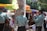 A group of musicians wearing light blue charro outfits with detailed embroidery are performing in an outdoor setting. They are playing guitars and standing near tables with people seated under colorful umbrellas. The scene appears lively and festive, with sunlight filtering through trees.