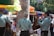 A group of musicians wearing light blue charro outfits with detailed embroidery are performing in an outdoor setting. They are playing guitars and standing near tables with people seated under colorful umbrellas. The scene appears lively and festive, with sunlight filtering through trees.
