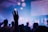 A dimly lit concert or event with a crowd of people. Hands are raised, and the scene is bathed in blue and purple lighting. Circular lights hang from the ceiling, creating a soft, atmospheric ambiance.