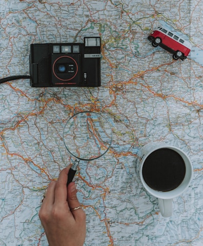 person holding magnifying glass beside black film camera white ceramic mug filled with black liquid near vehicle toy scale model
