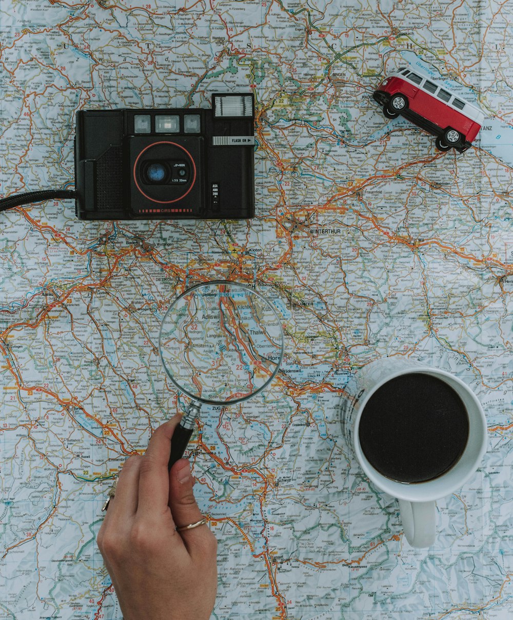 person holding magnifying glass beside black film camera white ceramic mug filled with black liquid near vehicle toy scale model