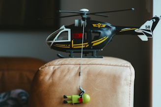 A toy helicopter is perched on a leather chair, with a small toy figure suspended underneath it by a string. The helicopter is black with yellow details and a white tail fin, marked with the number 58.