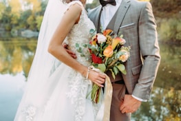 A bride in an embroidered white wedding dress holds a vibrant bouquet of flowers, while embracing a groom wearing a checked suit with a bow tie. They stand by a calm body of water with trees reflecting autumn colors in the background.
