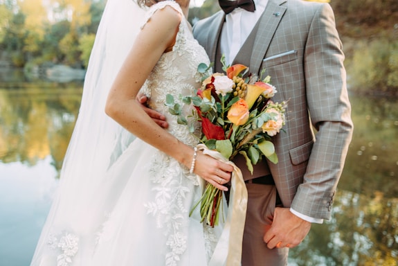 A bride in an embroidered white wedding dress holds a vibrant bouquet of flowers, while embracing a groom wearing a checked suit with a bow tie. They stand by a calm body of water with trees reflecting autumn colors in the background.