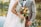 A bride in an embroidered white wedding dress holds a vibrant bouquet of flowers, while embracing a groom wearing a checked suit with a bow tie. They stand by a calm body of water with trees reflecting autumn colors in the background.