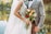 A bride in an embroidered white wedding dress holds a vibrant bouquet of flowers, while embracing a groom wearing a checked suit with a bow tie. They stand by a calm body of water with trees reflecting autumn colors in the background.