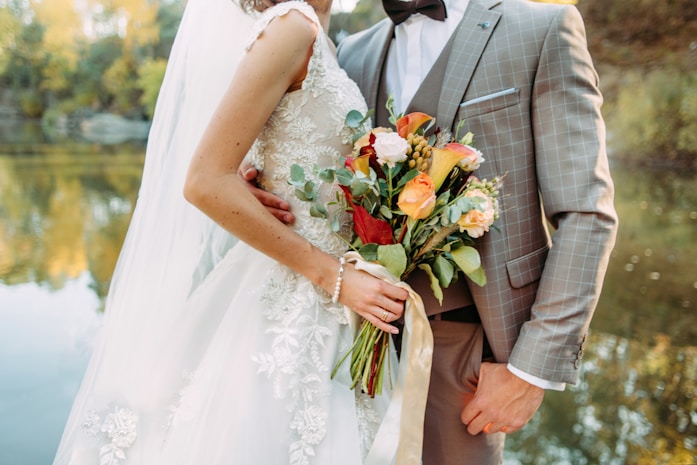 A bride in an embroidered white wedding dress holds a vibrant bouquet of flowers, while embracing a groom wearing a checked suit with a bow tie. They stand by a calm body of water with trees reflecting autumn colors in the background.