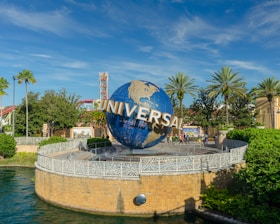 A large globe with the word 'Universal' prominently displayed, surrounded by lush greenery and palm trees. The scene is set in a theme park with a bright blue sky overhead. People can be seen walking near the globe, and there are structures and a body of water nearby.