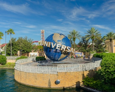 A large globe with the word 'Universal' prominently displayed, surrounded by lush greenery and palm trees. The scene is set in a theme park with a bright blue sky overhead. People can be seen walking near the globe, and there are structures and a body of water nearby.
