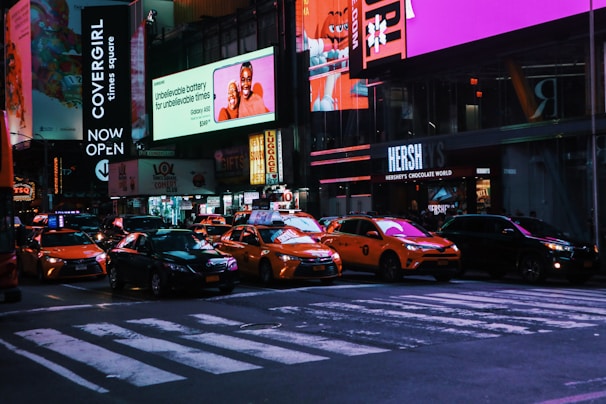A busy urban street at night with bright neon signs and billboards. Several yellow taxis and other cars are stopped in traffic. Large advertising screens display various brands and images, creating a lively and vibrant atmosphere. Pedestrians can be seen walking alongside the buildings that line the street.