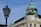 A historical building with intricate architectural details, featuring a rounded green copper dome and sculptures on its façade. The word 'GENERALI' is displayed on the dome. A classical street lamp is in the foreground, contrasted against a clear blue sky.