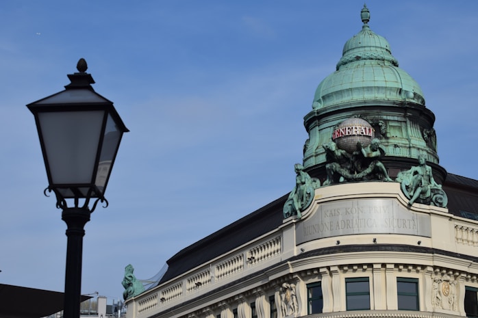 A historical building with intricate architectural details, featuring a rounded green copper dome and sculptures on its fa&ccedil;ade. The word 'GENERALI' is displayed on the dome. A classical street lamp is in the foreground, contrasted against a clear blue sky.