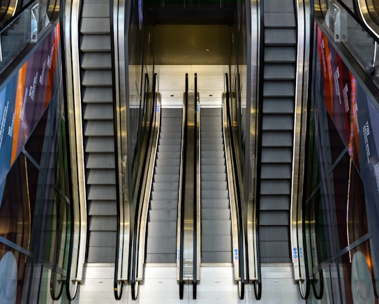 The image features a modern interior with a set of escalators and staircases. There are two staircases on either side with two parallel escalators in the middle. The escalators and stairs are surrounded by glass panels with advertisements and reflections visible. The lighting is soft, with a neutral color tone.