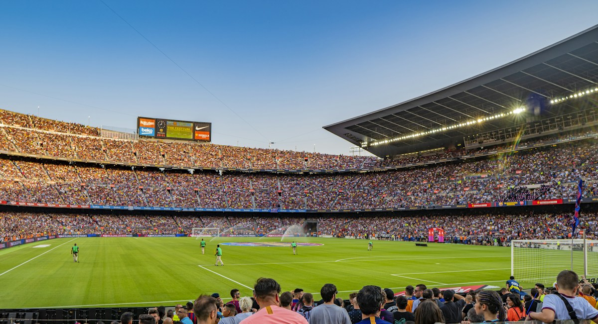 Des spectateurs suivent un match de football depuis les tribunes d’un stade en plein jour