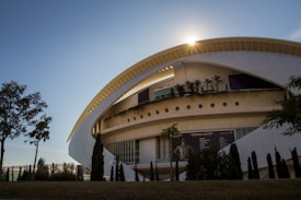 A modern, elliptical structure with a sleek design featuring arched elements and a sunlit backdrop. The building has a rooftop garden with small trees and plants lining the ledge. Tall windows adorn the facade, and various small trees or bushes are planted in the foreground, adding greenery to the scene. A banner with images and text is visible on the side of the building.