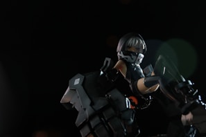 A stylized figure with mechanical attire, featuring a hood and mask. The character is in a dynamic pose, surrounded by dark, metallic elements that appear to be part of a robotic or futuristic environment. The lighting is dramatic, highlighting the figure against a dark background.
