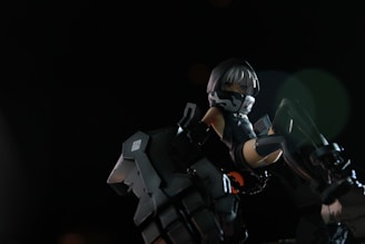 A stylized figure with mechanical attire, featuring a hood and mask. The character is in a dynamic pose, surrounded by dark, metallic elements that appear to be part of a robotic or futuristic environment. The lighting is dramatic, highlighting the figure against a dark background.
