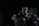 A stylized figure with mechanical attire, featuring a hood and mask. The character is in a dynamic pose, surrounded by dark, metallic elements that appear to be part of a robotic or futuristic environment. The lighting is dramatic, highlighting the figure against a dark background.