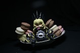 A small, anime-style figurine with blonde hair sits surrounded by colorful macarons on a white dish. The figure is dressed in dark, armor-like clothing with pointy elements, creating a playful contrast with the vibrant sweets. The scene is set against a dark background, drawing attention to the figure and the macarons.