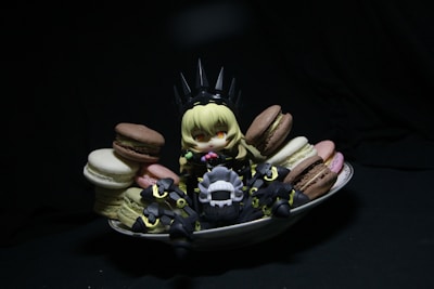 A small, anime-style figurine with blonde hair sits surrounded by colorful macarons on a white dish. The figure is dressed in dark, armor-like clothing with pointy elements, creating a playful contrast with the vibrant sweets. The scene is set against a dark background, drawing attention to the figure and the macarons.