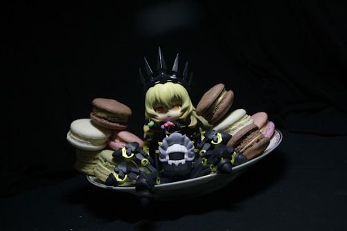 A small, anime-style figurine with blonde hair sits surrounded by colorful macarons on a white dish. The figure is dressed in dark, armor-like clothing with pointy elements, creating a playful contrast with the vibrant sweets. The scene is set against a dark background, drawing attention to the figure and the macarons.