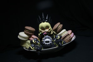 A small, anime-style figurine with blonde hair sits surrounded by colorful macarons on a white dish. The figure is dressed in dark, armor-like clothing with pointy elements, creating a playful contrast with the vibrant sweets. The scene is set against a dark background, drawing attention to the figure and the macarons.