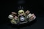 A small, anime-style figurine with blonde hair sits surrounded by colorful macarons on a white dish. The figure is dressed in dark, armor-like clothing with pointy elements, creating a playful contrast with the vibrant sweets. The scene is set against a dark background, drawing attention to the figure and the macarons.