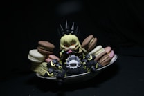 A small, anime-style figurine with blonde hair sits surrounded by colorful macarons on a white dish. The figure is dressed in dark, armor-like clothing with pointy elements, creating a playful contrast with the vibrant sweets. The scene is set against a dark background, drawing attention to the figure and the macarons.