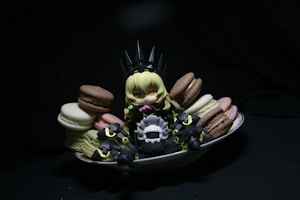 A small, anime-style figurine with blonde hair sits surrounded by colorful macarons on a white dish. The figure is dressed in dark, armor-like clothing with pointy elements, creating a playful contrast with the vibrant sweets. The scene is set against a dark background, drawing attention to the figure and the macarons.