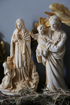 Two religious statues are displayed, depicting figures in a traditional style. One statue shows a figure with a long robe and hands held together in prayer, while the other statue includes a bearded figure holding a child. Both are surrounded by decorative elements and positioned against a neutral background.