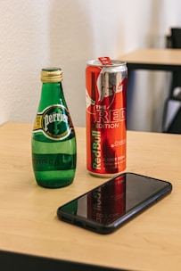 A green glass bottle of Perrier sparkling water and a red can of Red Bull energy drink are placed on a light wooden table. An unopened black smartphone rests horizontally in front of the drinks.