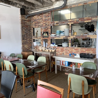 A cozy restaurant interior with a rustic theme, featuring brick walls and a visible kitchen area. The dining area has tables with mismatched chairs, set with glasses and napkins. The shelves display stacked bowls and plates, and hanging salami adds a touch of authenticity. The ceiling shows exposed pipes and industrial elements.