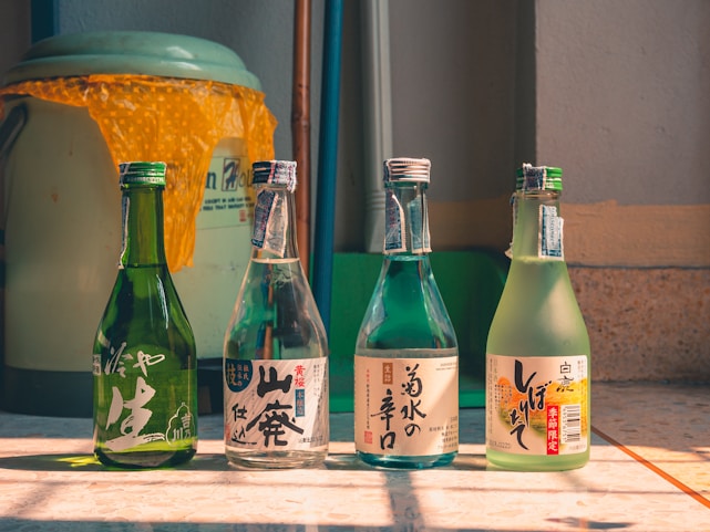 Four bottles of sake are placed on a sunlit surface with Japanese labels on them, each showcasing unique designs and colors. They are arranged in a row with a small gap between each. In the background, a large container is partially visible, draped with an orange textured cover.