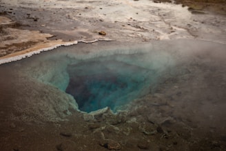A geothermal hot spring pool surrounded by rocky terrain, with steam rising from the surface. The pool has a deep, vibrant blue color at its center, transitioning to lighter shades as it reaches the edges. The landscape around it is barren, with mineral deposits visible along the edges of the pool.