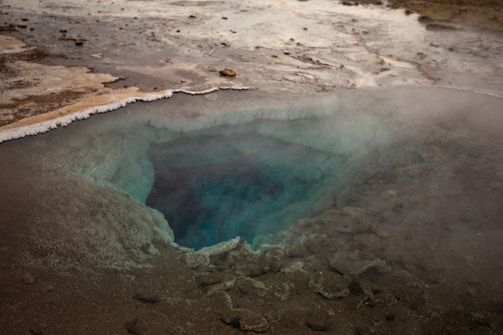 A geothermal hot spring pool surrounded by rocky terrain, with steam rising from the surface. The pool has a deep, vibrant blue color at its center, transitioning to lighter shades as it reaches the edges. The landscape around it is barren, with mineral deposits visible along the edges of the pool.