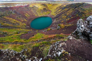 A scenic view of a volcanic crater filled with vibrant turquoise water, surrounded by multicolored rocky and mossy terrain. The landscape is dominated by earthy tones with lush green patches, creating a striking contrast with the vivid blue of the crater lake. The rocky cliffs are rugged with various textures, adding depth to the scene.