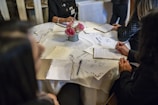 A group of people sitting around a round table with notebooks and menus in front of them. The table is covered with a white tablecloth, and there is a small bouquet of pink flowers in the center. The individuals are engaged in writing or taking notes, suggesting a formal or educational setting.