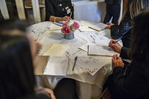 A group of people sitting around a round table with notebooks and menus in front of them. The table is covered with a white tablecloth, and there is a small bouquet of pink flowers in the center. The individuals are engaged in writing or taking notes, suggesting a formal or educational setting.