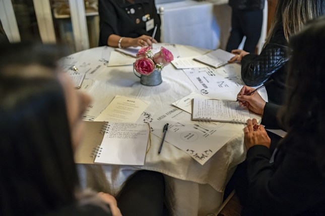 A group of people sitting around a round table with notebooks and menus in front of them. The table is covered with a white tablecloth, and there is a small bouquet of pink flowers in the center. The individuals are engaged in writing or taking notes, suggesting a formal or educational setting.