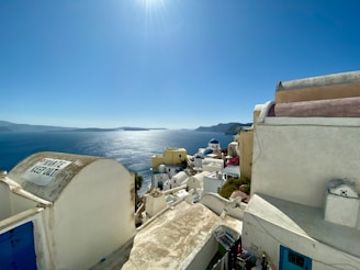 A scenic view of white buildings with blue domes on a hillside overlooking a shimmering blue sea. The sky is clear and bright, with the sun casting strong light across the landscape. A 'Private Keep Out' sign is visible on the foreground structure.