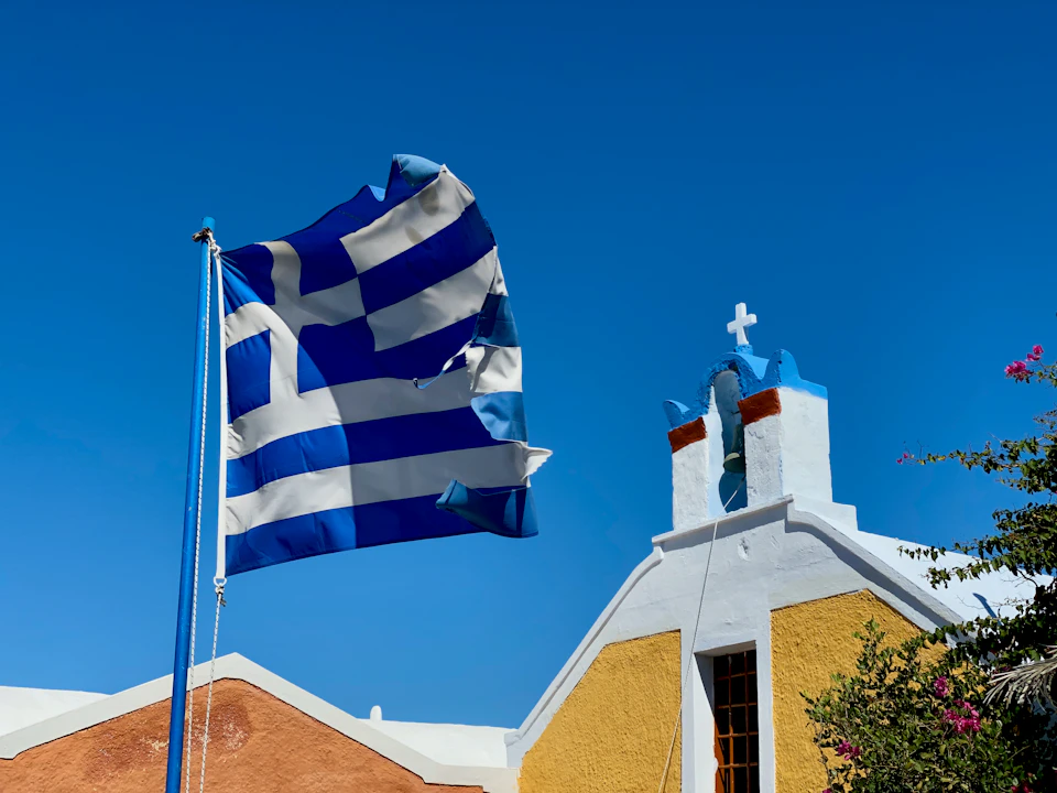 Greek flag flies high on pole amidst traditional church in clear blue sky background