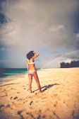 A person in swimwear stands on a sandy beach, facing the ocean with a hand on their head. The sky is partly cloudy with a faint rainbow visible near the horizon. The beach is expansive, with footprints visible in the sand.