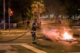 A firefighter in protective gear is standing on a street holding a hose, facing a small fire burning on the ground. The fire is surrounded by metal barricades, and it is nighttime with the scene illuminated by streetlights. Several people can be seen in the background, some walking and others standing, observing the situation.