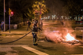 A firefighter in protective gear is standing on a street holding a hose, facing a small fire burning on the ground. The fire is surrounded by metal barricades, and it is nighttime with the scene illuminated by streetlights. Several people can be seen in the background, some walking and others standing, observing the situation.